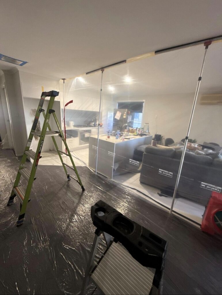 Floor-to-ceiling poly containment wall with spring-loaded poles separating the work zone from the kitchen and lounge